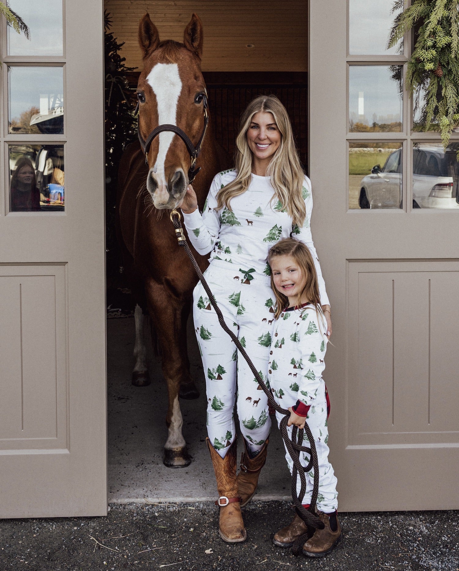 Woman and child in matching pajamas standing next to a horse in a stable.