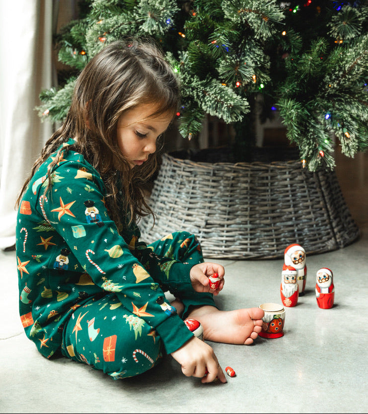 Child in green pajamas sitting on the floor next to a Christmas tree with small Russian dolls.