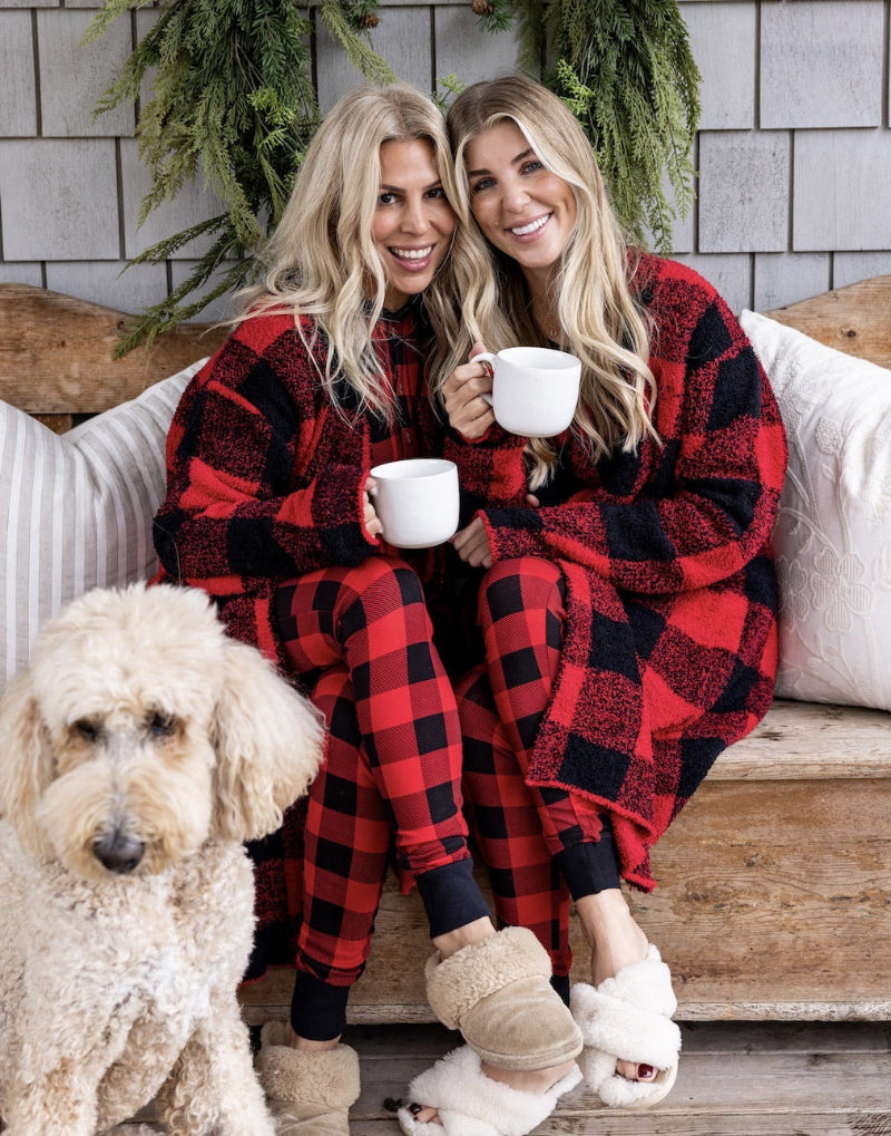 Two women in red and black plaid pajamas sitting on a wooden bench with a dog, holding mugs.