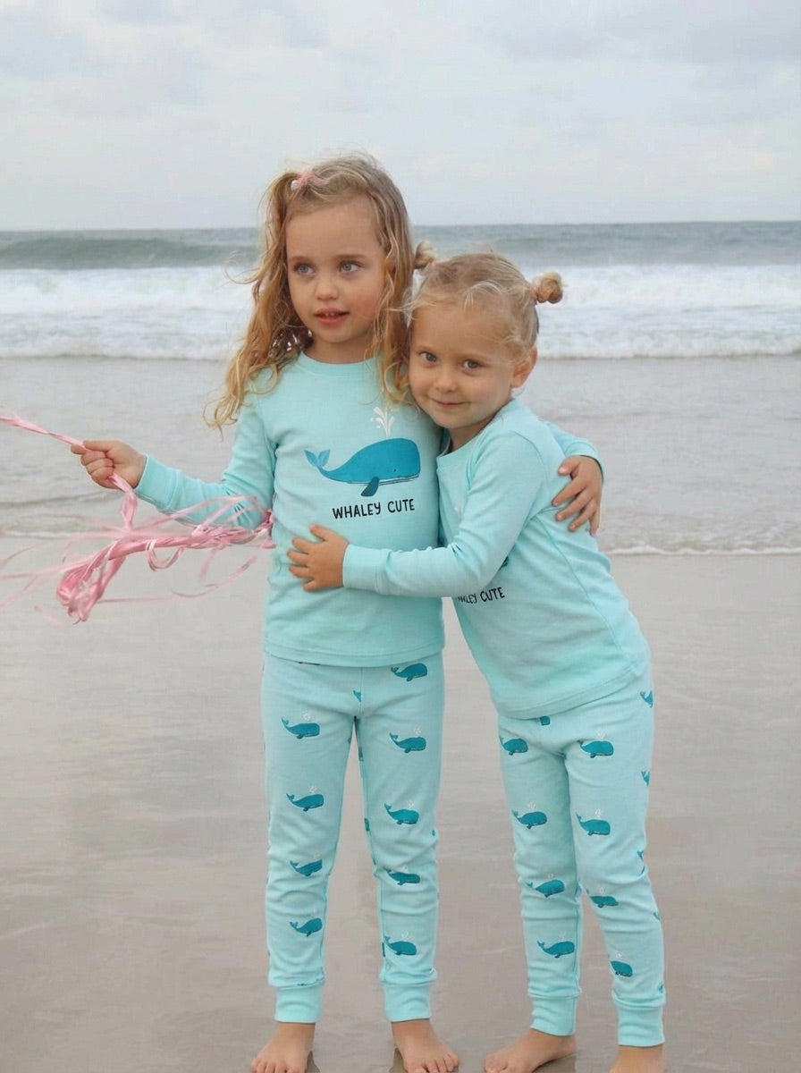 Two children in matching light blue pajamas with whale patterns on a beach.