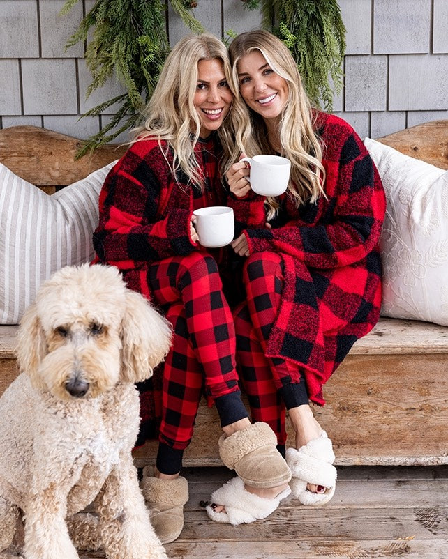 Two women in red and black plaid pajamas sitting on a wooden bench with a dog.