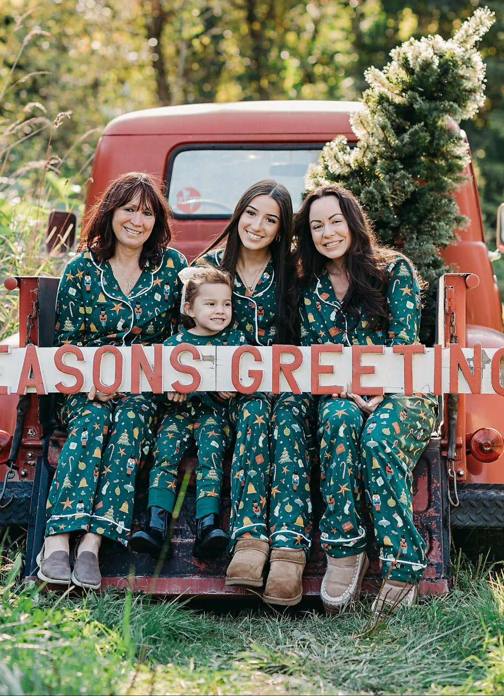 Three women and a child sitting in the back of a red truck with a 'Season's Greetings' sign, surrounded by trees.