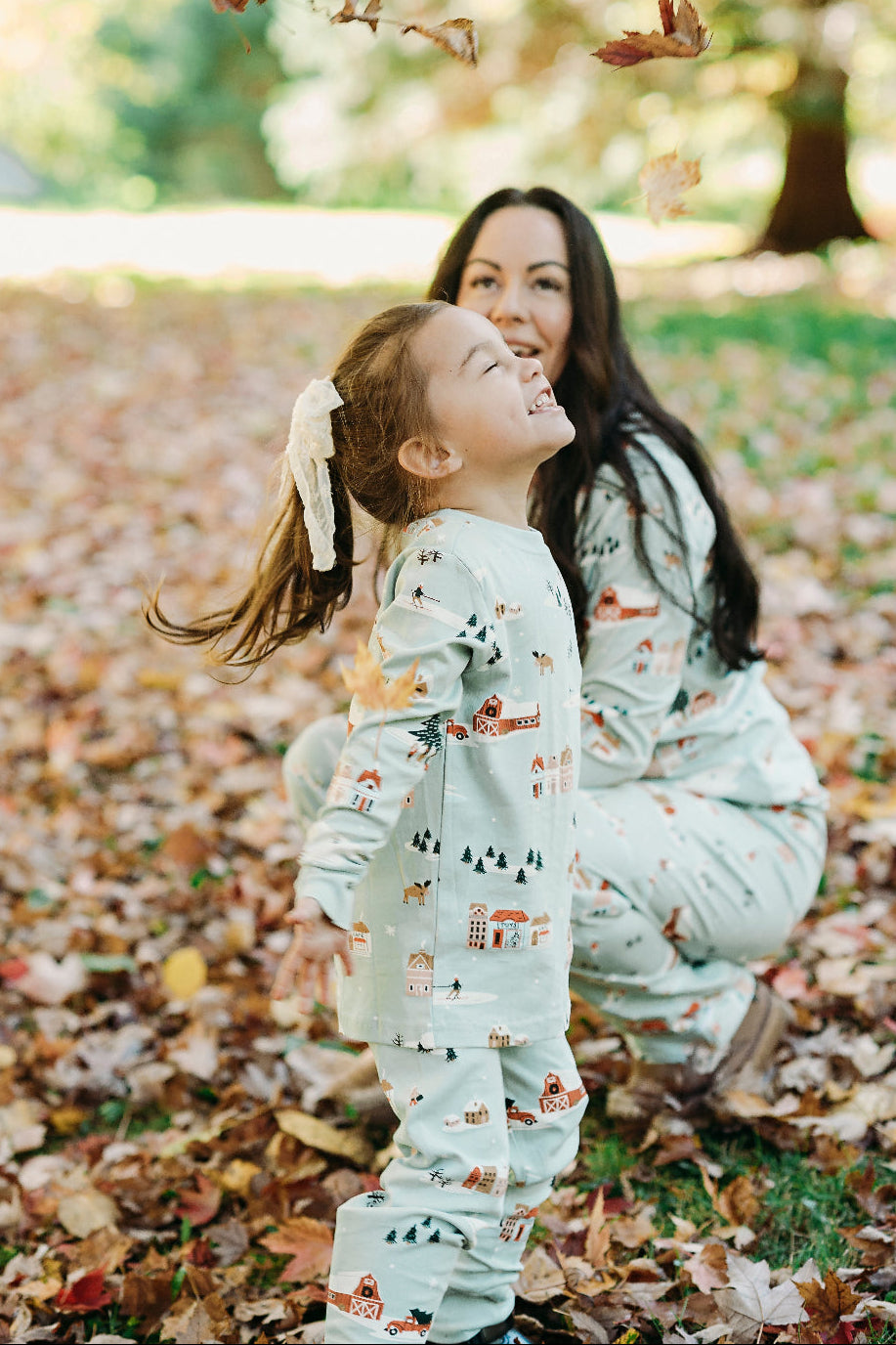 Mother and daughter in matching pajamas playing in a park with falling leaves.