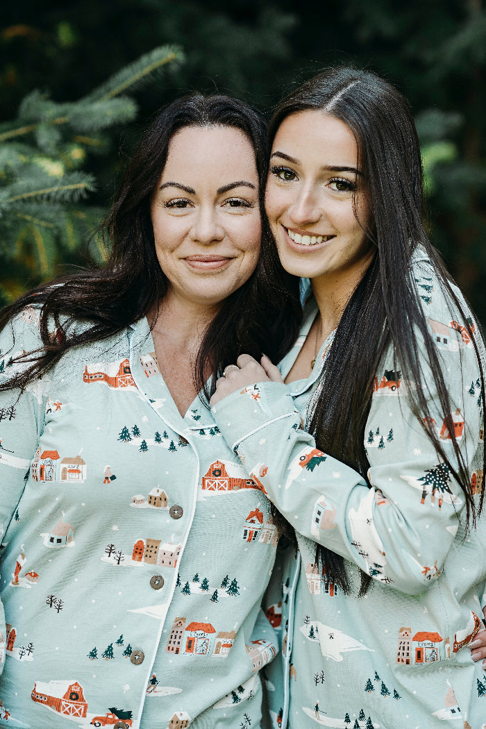 Two women wearing matching pajama sets with a winter village pattern, standing close together outdoors.