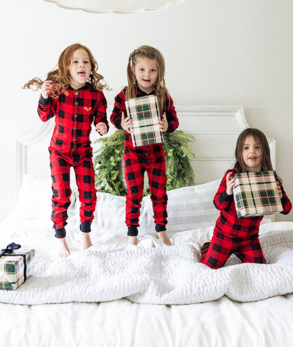 Three children in red and black plaid pajamas standing on a bed with Christmas decorations.