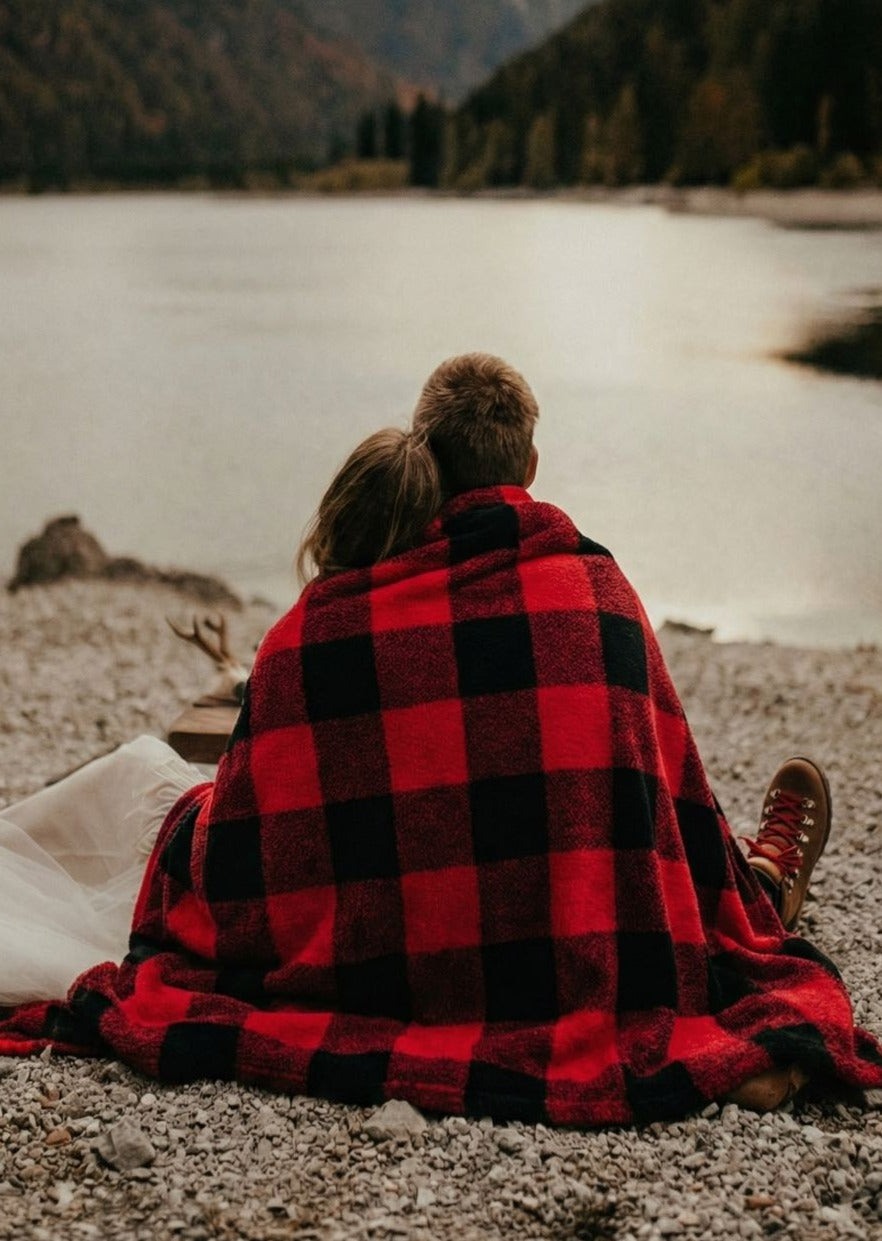 Two people sitting together on a rocky shore wrapped in a red and black checkered blanket with a scenic lake and mountains in the background.